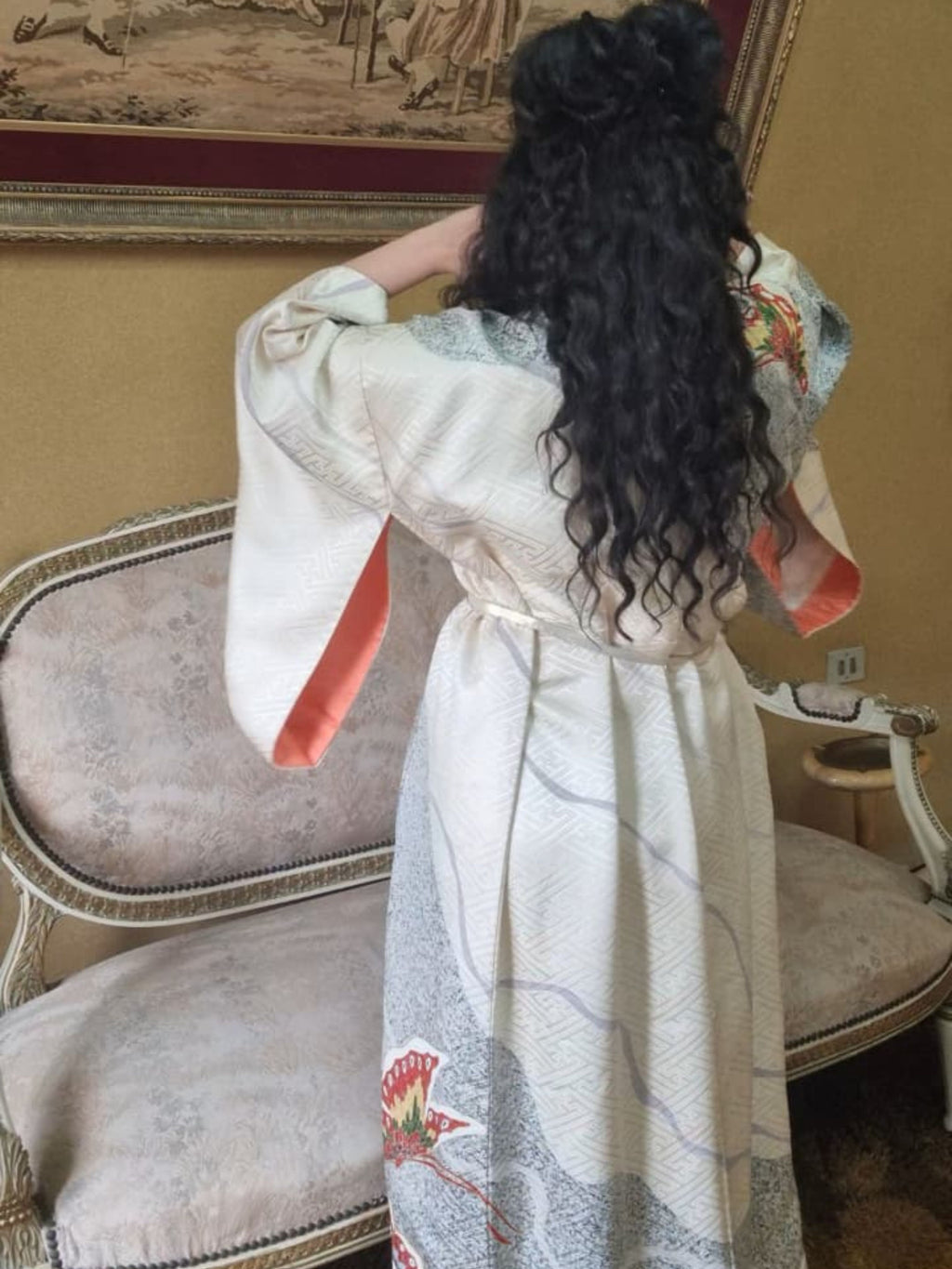 Woman in a floral kimono standing in a room with a decorative wall and chair.