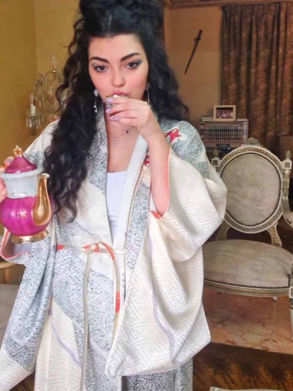 Woman in a patterned shawl holding a teapot in an ornate room.