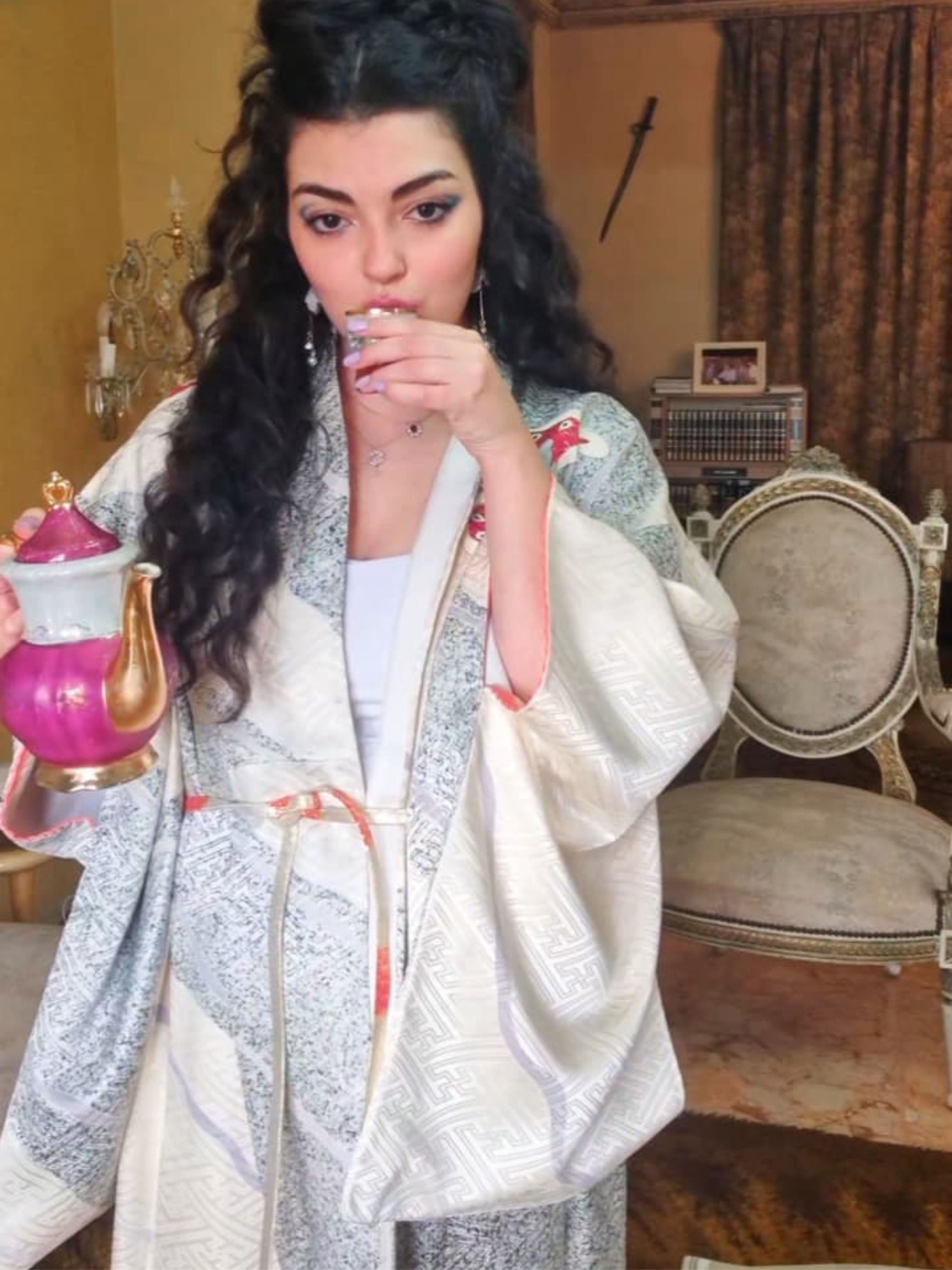 Woman in a patterned shawl holding a teapot in an ornate room.