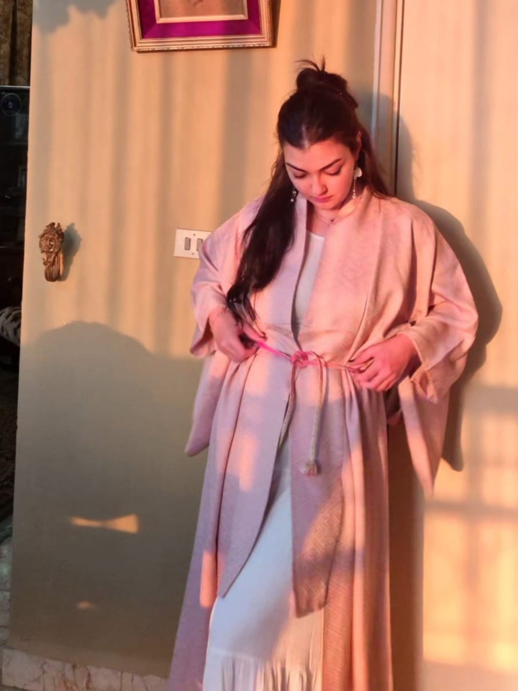 Woman wearing a light pink traditional Japanese kimono adjusting the pink obi belt in front of a mirror with warm lighting.