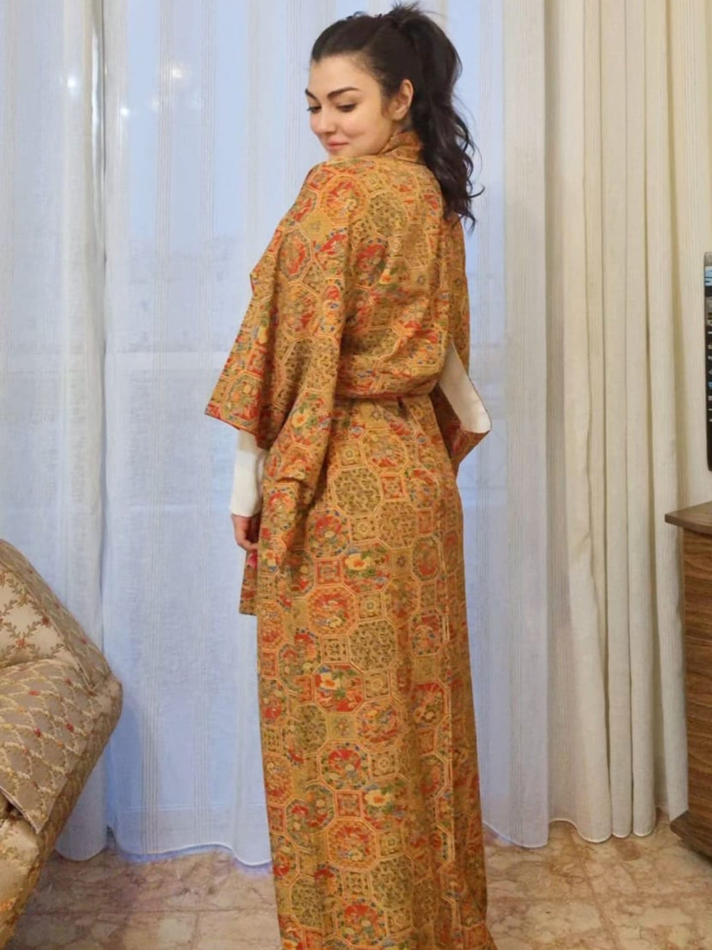 Woman wearing a long, patterned dress standing in a room with curtains and a couch.