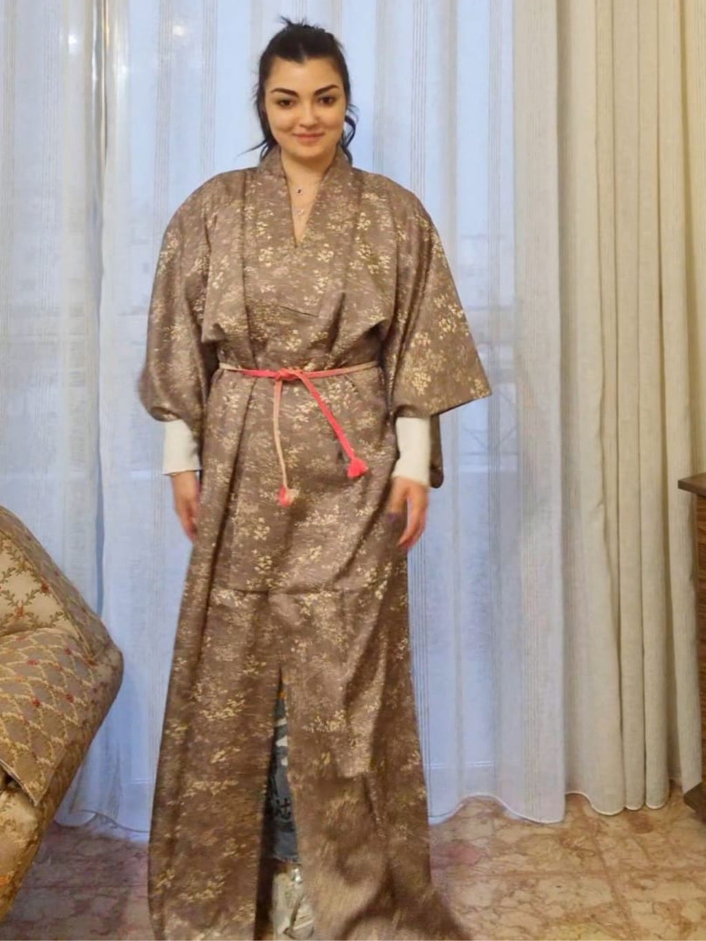Woman wearing a Japanese grey patterned kimono with a belt in a room with curtains and furniture.