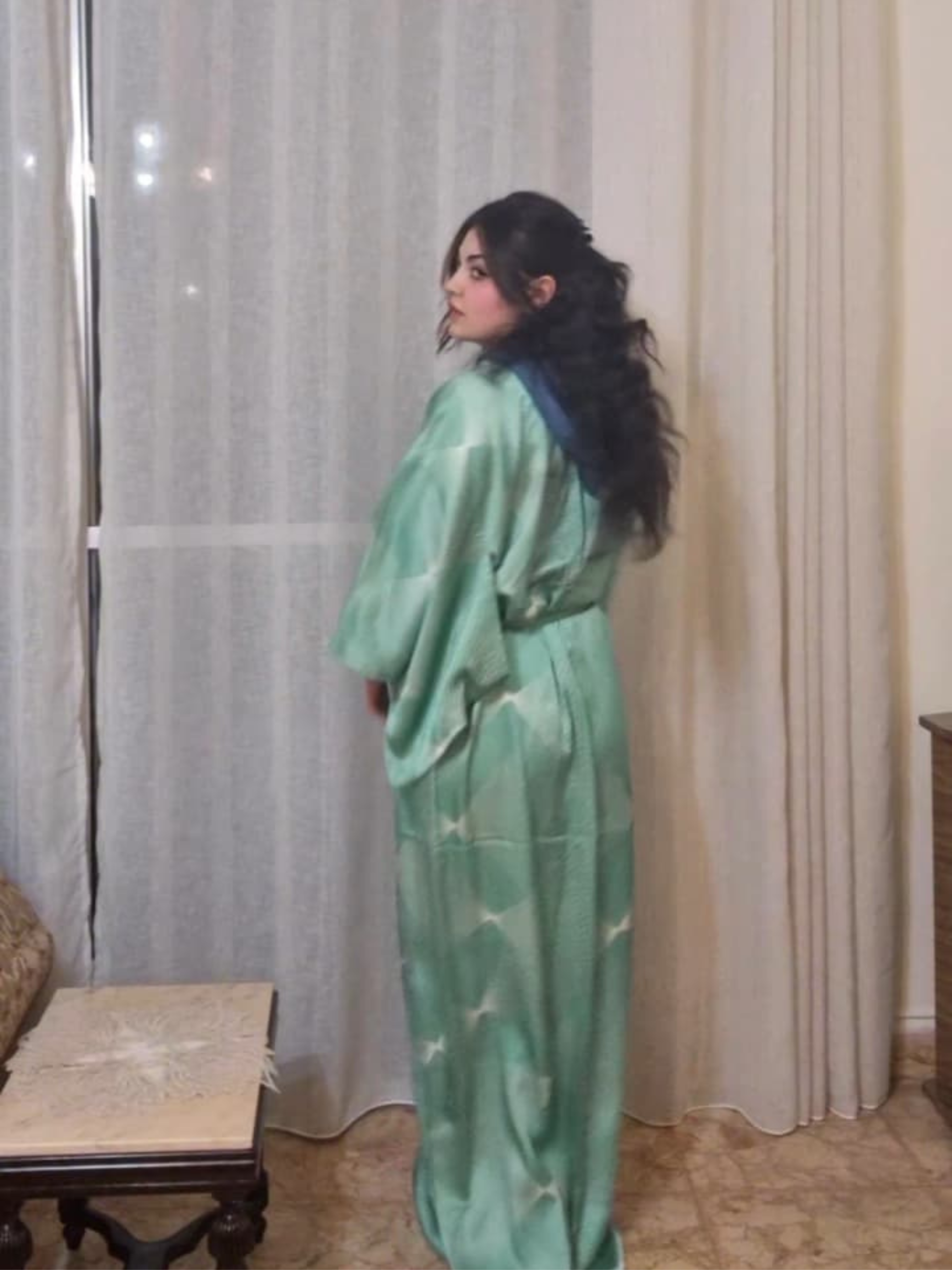 Woman wearing a green patterned dress standing in a room with curtains.