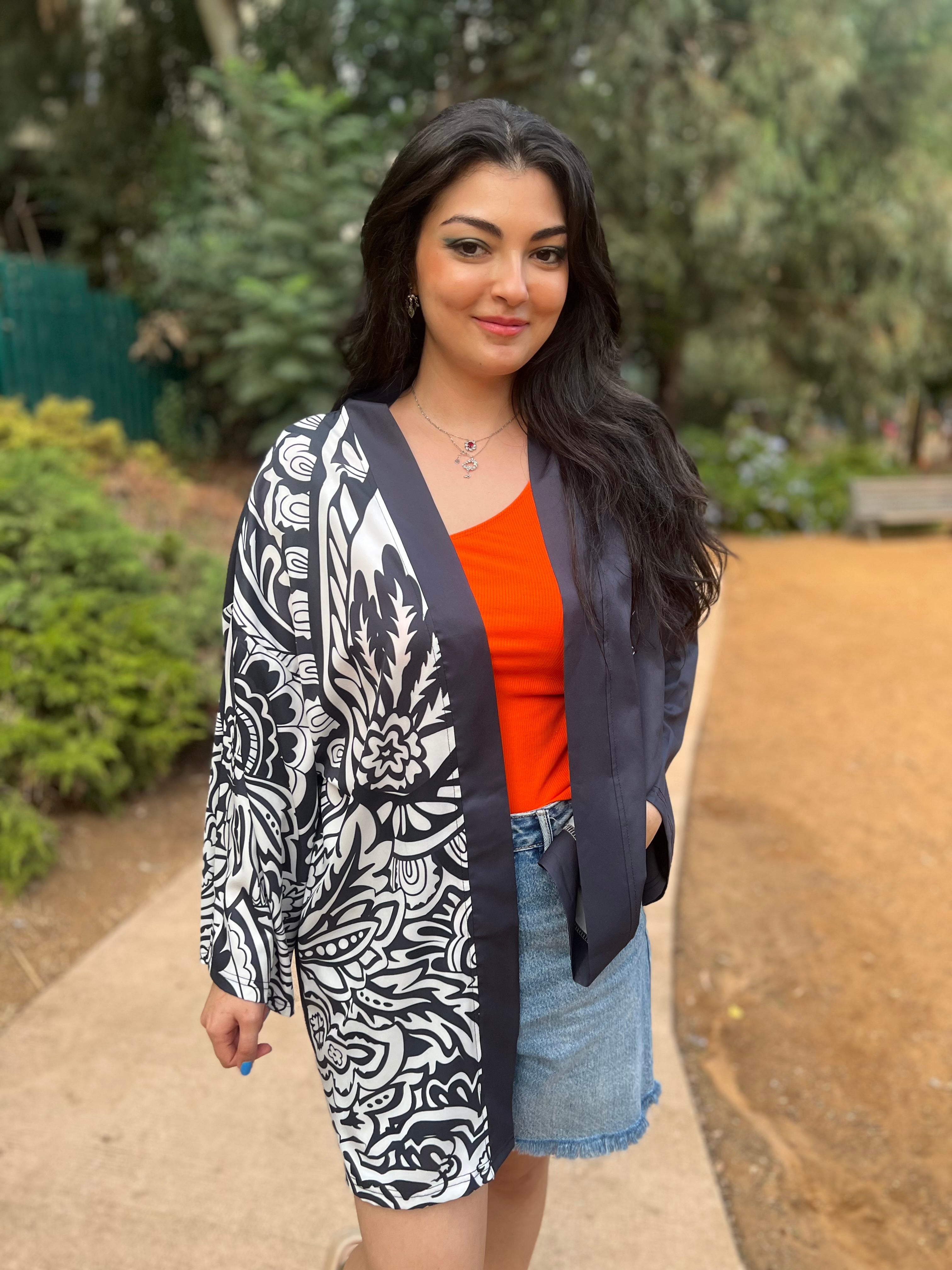 Woman wearing a black and white patterned cardigan over an orange top and denim shorts, standing outdoors.