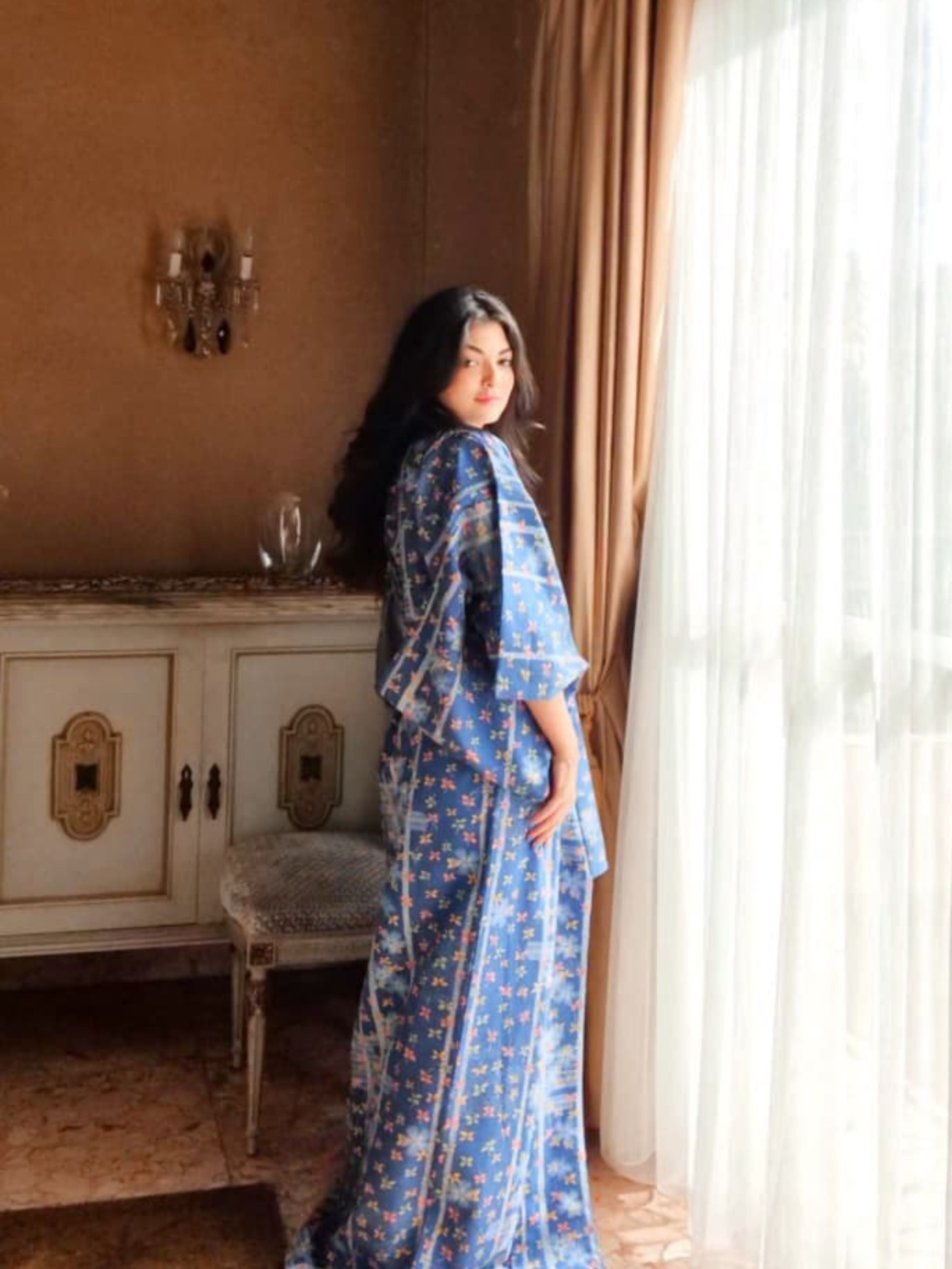 Woman in a blue floral kimono standing in a room with a window and decorative wall.