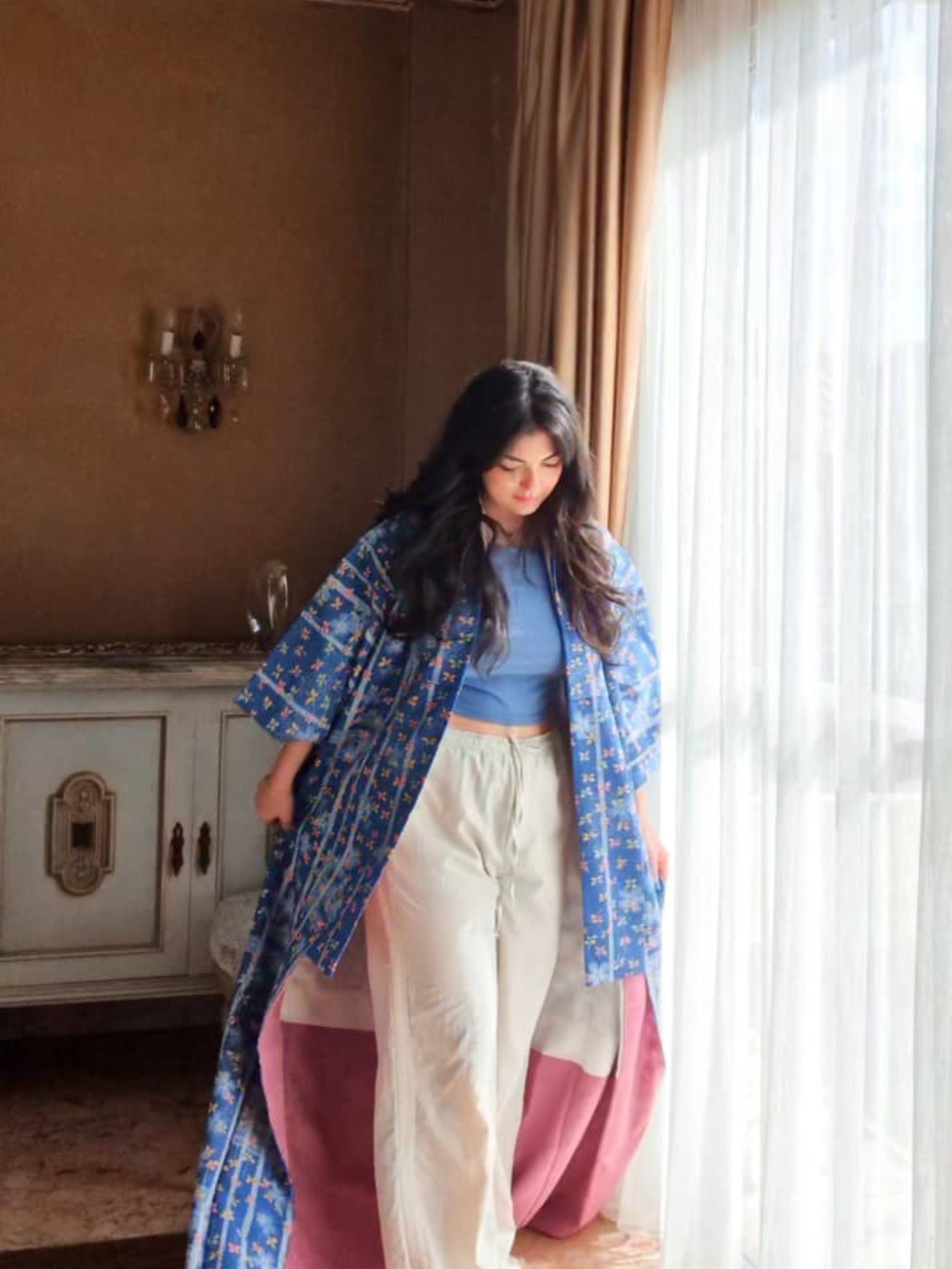 Woman wearing a blue floral kimono white pants standing in a room with a window and decorative wall.
