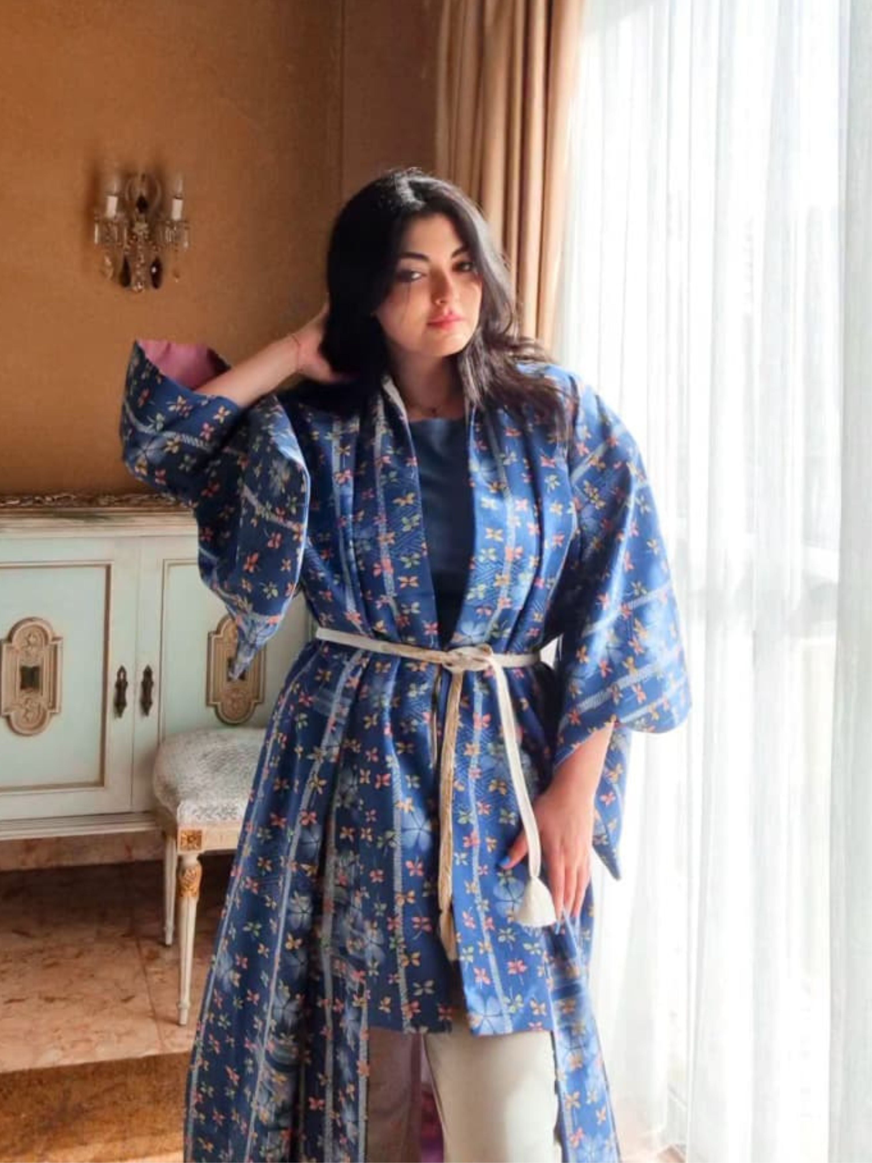 Woman wearing a blue floral kimono with obi belt in a room with a window and decorative elements.