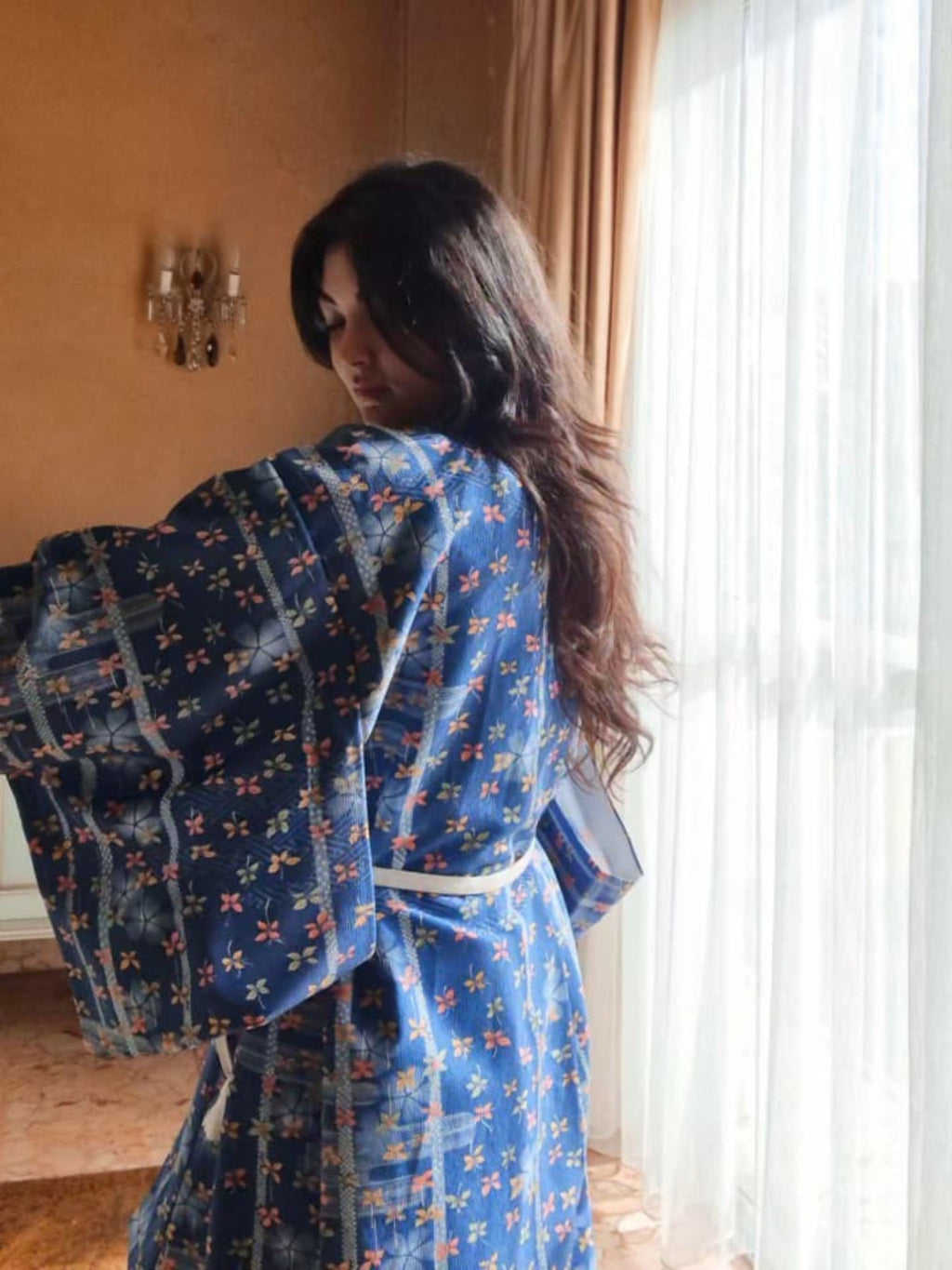 Woman wearing a blue floral kimono standing in a room with a window and wall decor.