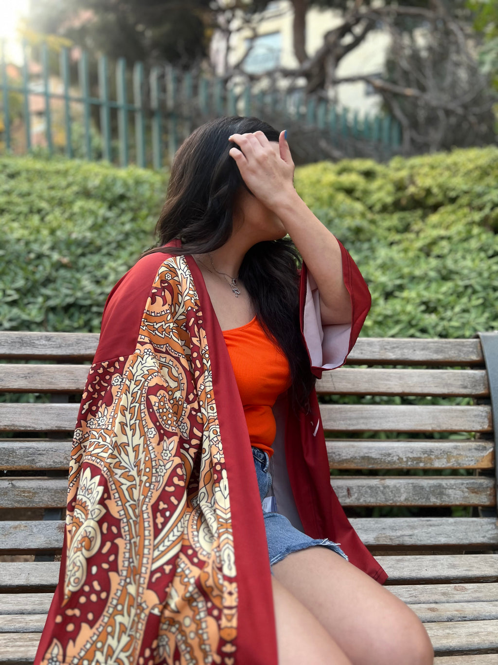 Woman sitting on a bench wearing an orange top and patterned red shawl in an outdoor setting.