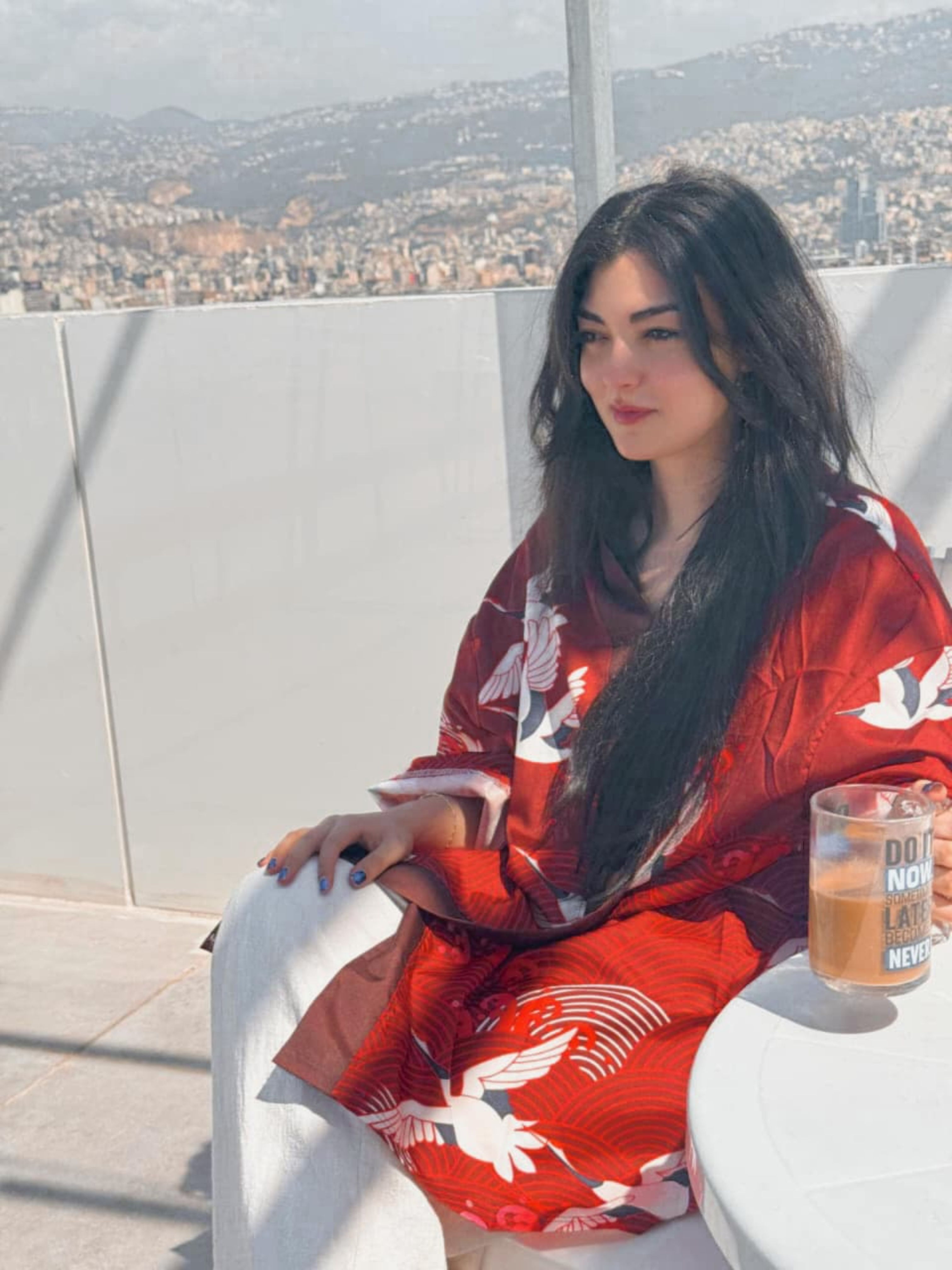 Woman in a red kimono-style outfit sitting on a rooftop with a cityscape in the background